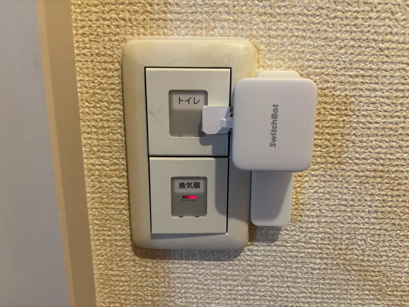 トイレの壁スイッチに取り付けたボット。表示付きの2連スイッチの上段(トイレ照明)をSmartBASE取付台で固定したボットが担当している