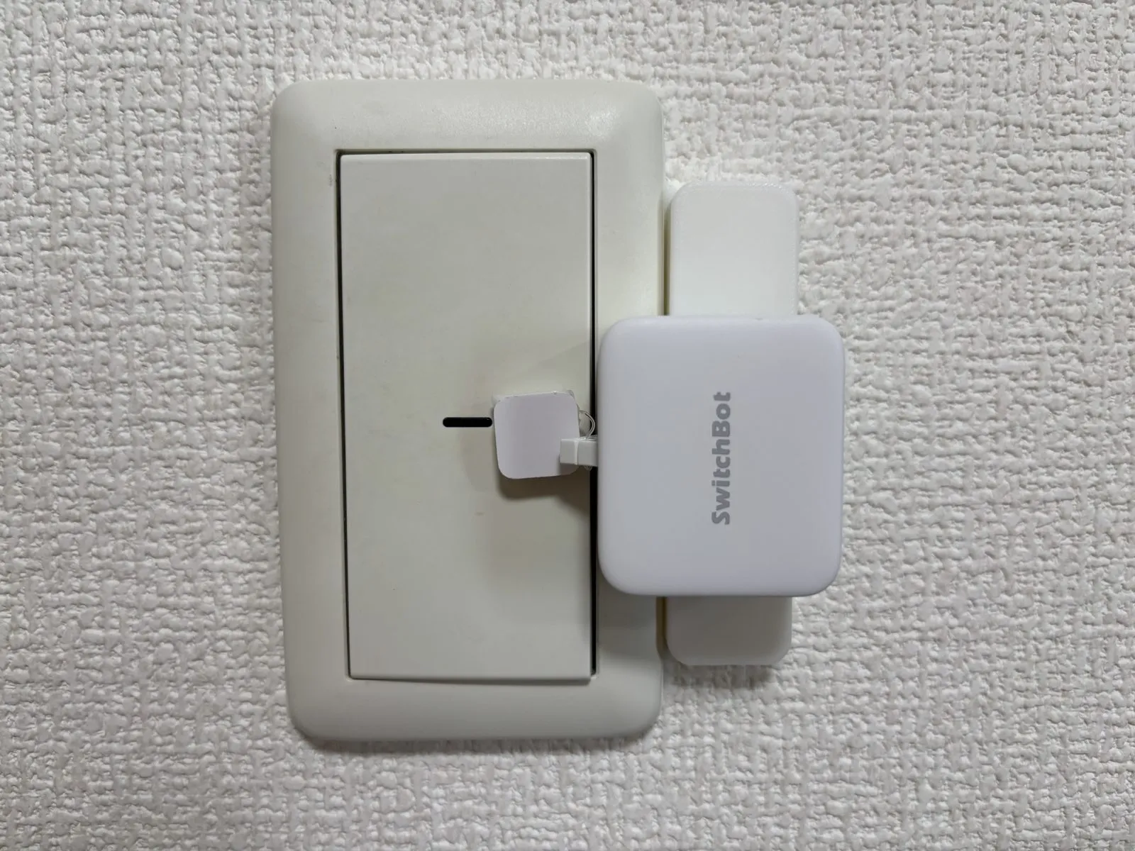 洗面所の壁スイッチに取り付けたSwitchBotボット。奥の縦長の白い台座がSmartBASE取付台、スイッチ面の白いタブがスイッチモード用のON/OFF用シート